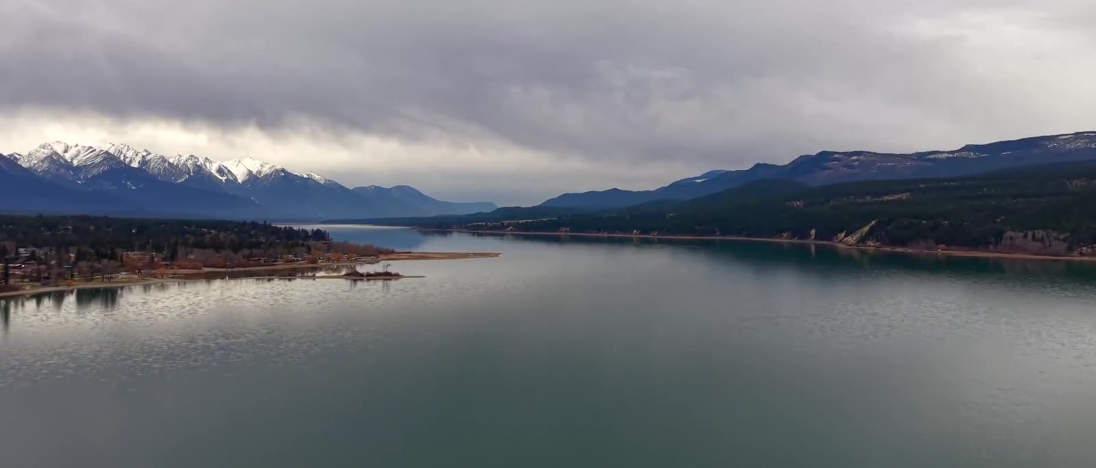 Aerial view of a mountain reservoir, a forested spit of shoreline and a lakeside community, with snow-capped Rockies on the horizon under heavy cloud.